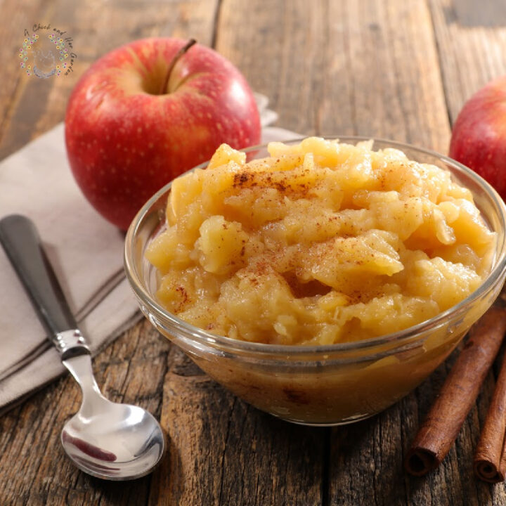 cinnamon applesauce in a glass dish on a wooden table with two apples, a spoon, napkin and cinnamon sticks