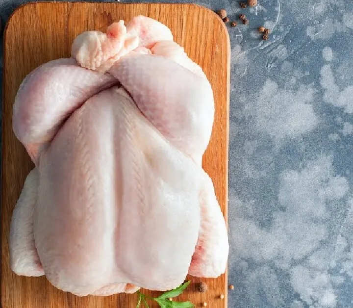 raw whole chicken on a wooden cutting board with jars of salt cloves of garlic herbs and peppercorns on concrete countertop