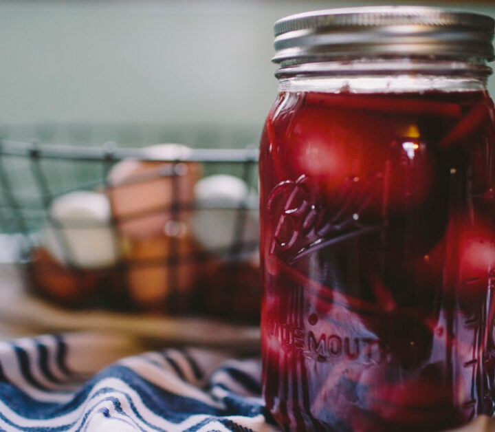 mason jar of pickled beet eggs on a blue an white towel with a wire basket of eggs in the background