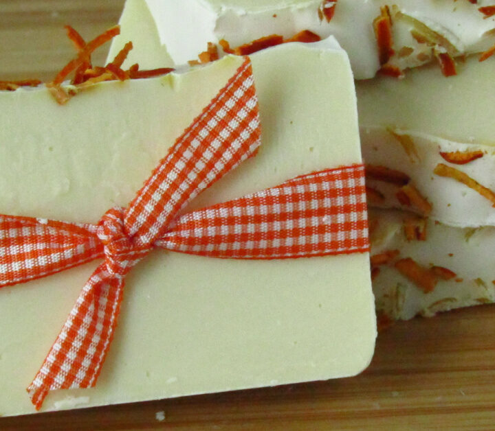 one bar of white soap with dried orange peels on top wrapped in orange gingham ribbon leaning against two stacked bars of soap