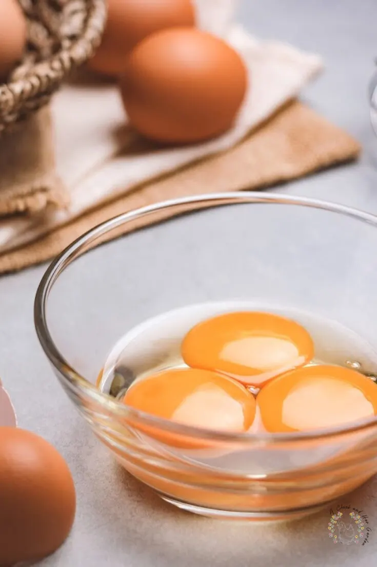 small glass dish holding three egg yolks with eggs on a dish towel in the background