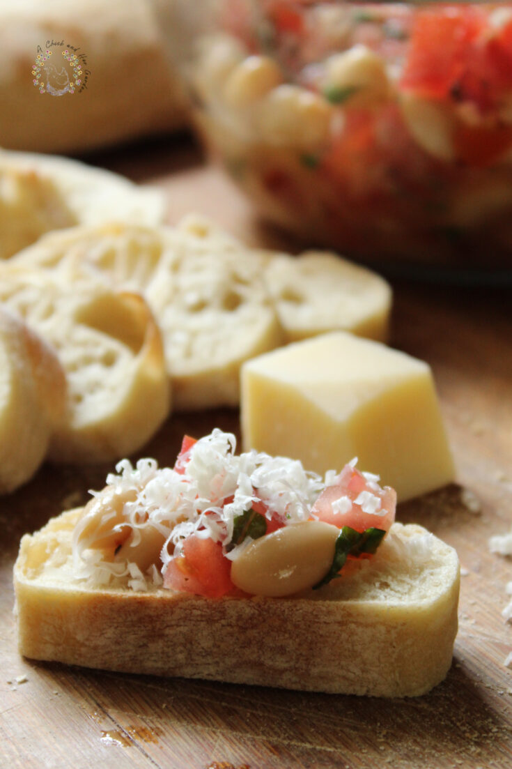 fresh bruschetta with grated parmesan on a slice of baguette with more baguette slices, a cube of fresh parmesan and a bowl of bruschetta mixture in the background