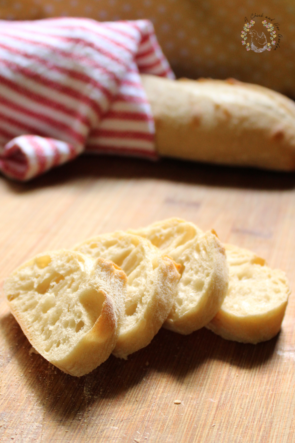 four slices of baguette on a wooden cutting board with a whole baguette wrapped in a redand white cloth in the background