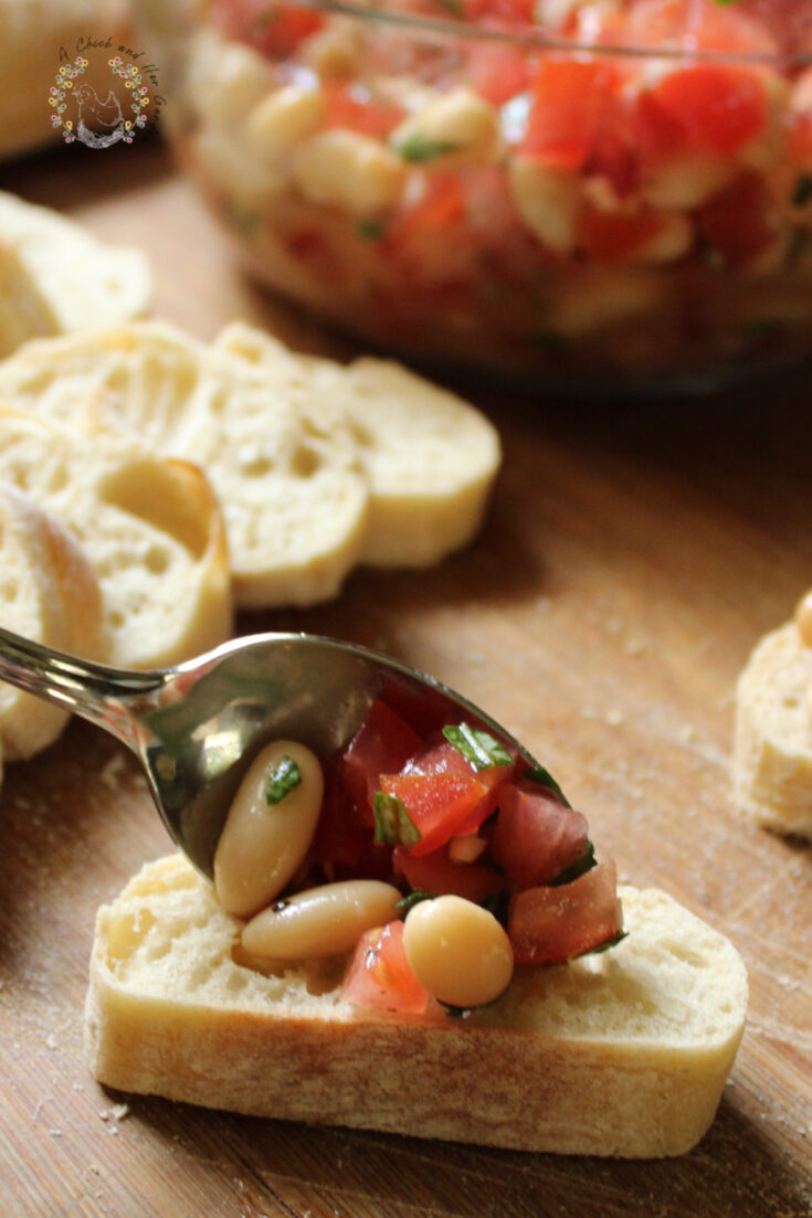 spoon placing bruschetta mixture onto a baguette slice with slices of baguette and bowl of bruschetta mixture in the background